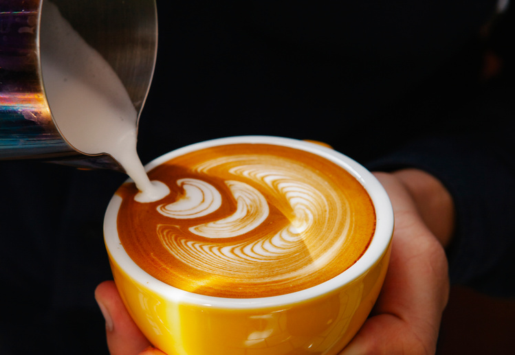 Crop barista pouring milk into coffee with foam in cafe